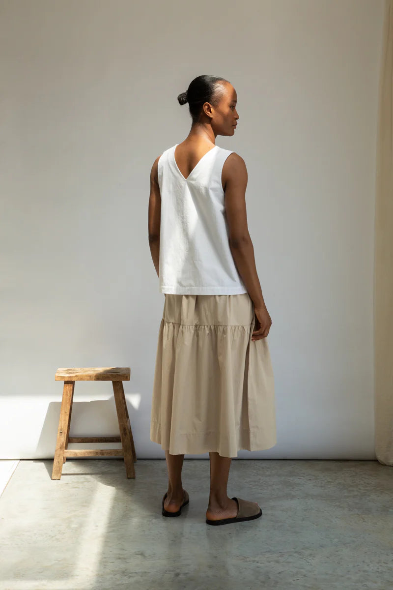 Woman wearing a white sleeveless top and beige skirt standing against a plain wall.