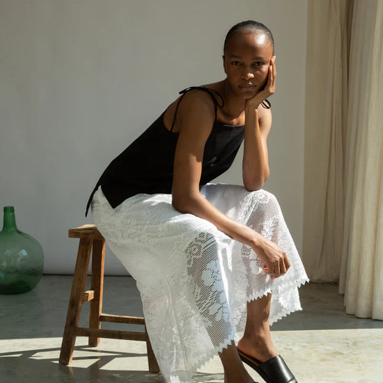 Woman sitting on a wooden stool wearing a black top and white lace Beaumont Organic skirt in a softly lit room.