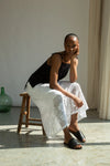 Woman sitting on a wooden stool wearing a black top and white lace Beaumont Organic skirt in a softly lit room.