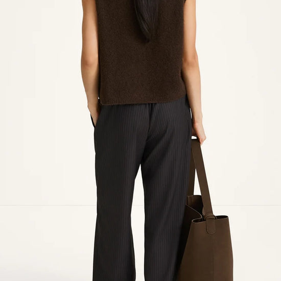 Person wearing a brown sleeveless top and black pants, holding a brown tote bag on a white background