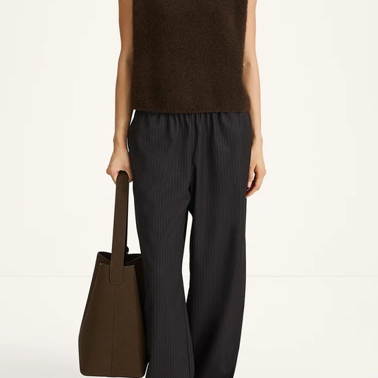 A model wearing a sleeveless dark brown knit mock-neck top and dark charcoal gray pinstriped wide-leg trousers, holding a dark brown tote bag against a clean white studio background.