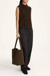 Person wearing a brown sleeveless sweater and black pants, holding a matching brown tote bag on a white background.