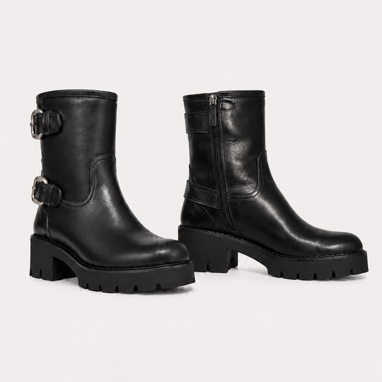 A pair of black leather ankle boots with chunky lug soles and buckle straps on the side, displayed against a light gray studio background.