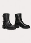 A pair of black leather ankle boots with chunky lug soles and buckle straps on the side, displayed against a light gray studio background.