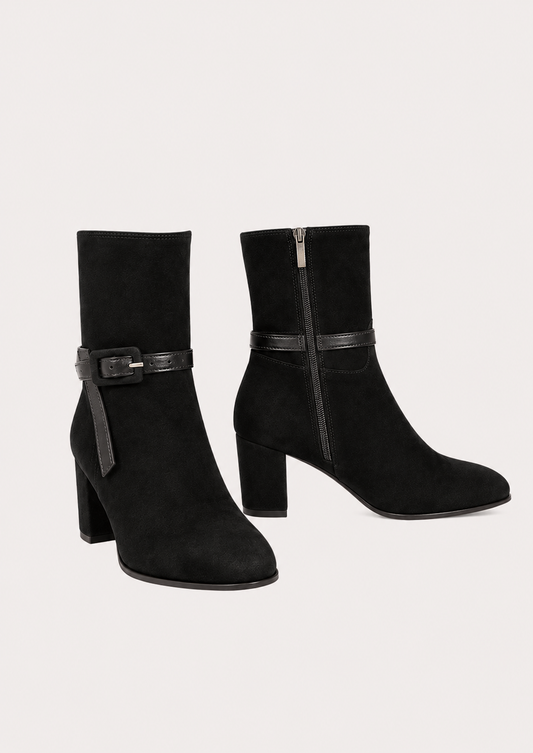 A pair of black suede ankle boots with block heels and a decorative strap detail across the ankle, shown against a light beige background.