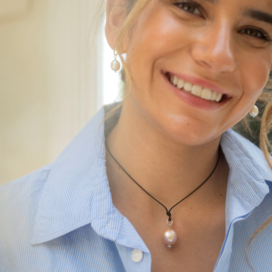 Woman wearing a light blue shirt and a necklace with a pink pendant, smiling outdoors.