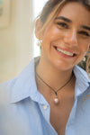 Woman wearing a light blue shirt and a necklace with a pink pendant, smiling outdoors.