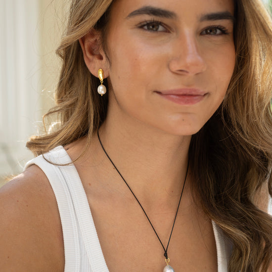 Woman wearing a white tank top and pearl necklace with earrings, outdoors.