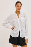 Woman wearing a white blouse with small colorful patterns and black shorts on a light background