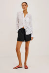 Woman wearing a white blouse with black patterns and black shorts on a light gray background