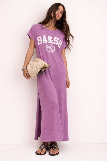 Woman wearing a purple dress with 'BA&SH' print, holding a beige bag against a white background