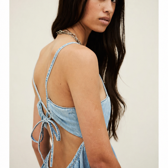 Woman wearing a light blue denim dress with thin straps and open back against a plain background