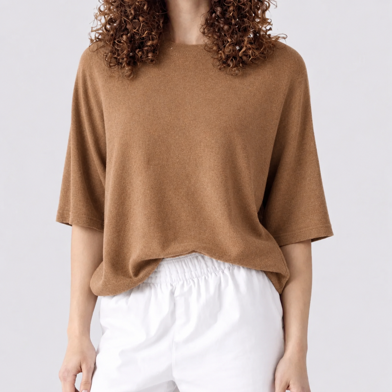 A person wearing a loose, short-sleeve knit top in a warm camel brown color, paired with white drawstring pants, against a plain light gray studio background.