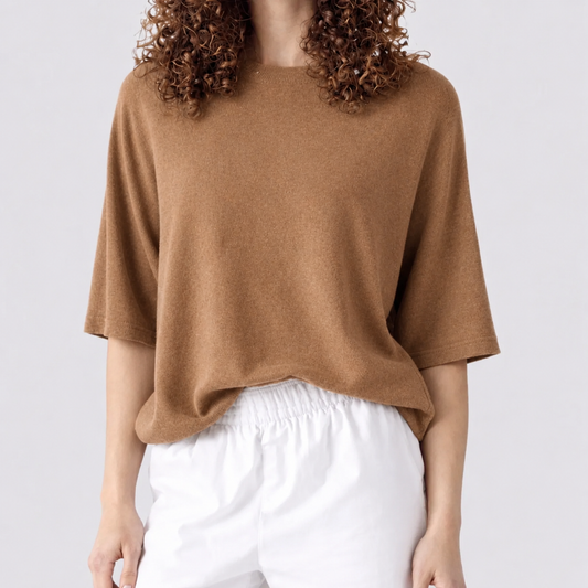 A person wearing a loose, short-sleeve knit top in a warm camel brown color, paired with white drawstring pants, against a plain light gray studio background.