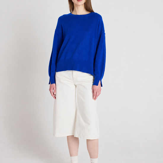 A woman stands against a light gray studio background wearing a bright royal blue knit sweater, off-white cropped shorts, white socks, and dark brown sneakers.