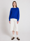 A woman stands against a light gray studio background wearing a bright royal blue knit sweater, off-white cropped shorts, white socks, and dark brown sneakers.