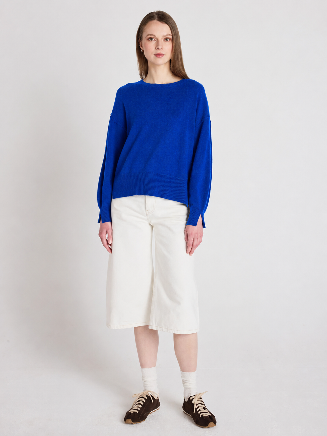 A woman stands against a light gray studio background wearing a bright royal blue knit sweater, off-white cropped shorts, white socks, and dark brown sneakers.