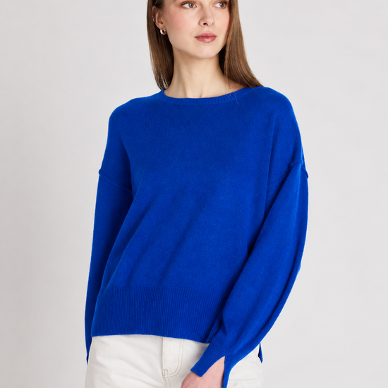 A woman wearing a vibrant royal blue knit sweater with a boat neckline and wide, slightly dropped shoulders, paired with white pants, set against a plain light gray studio background.