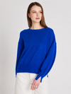 A woman wearing a vibrant royal blue knit sweater with a boat neckline and wide, slightly dropped shoulders, paired with white pants, set against a plain light gray studio background.