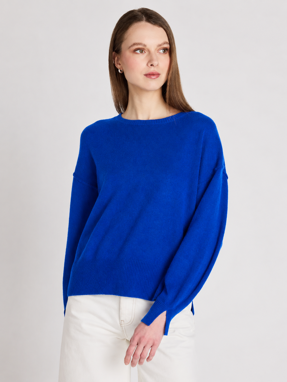 A woman wearing a vibrant royal blue knit sweater with a boat neckline and wide, slightly dropped shoulders, paired with white pants, set against a plain light gray studio background.
