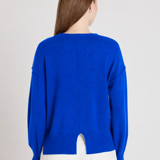 A woman viewed from the back wearing a bright royal blue knit sweater and white pants against a plain light gray studio background.