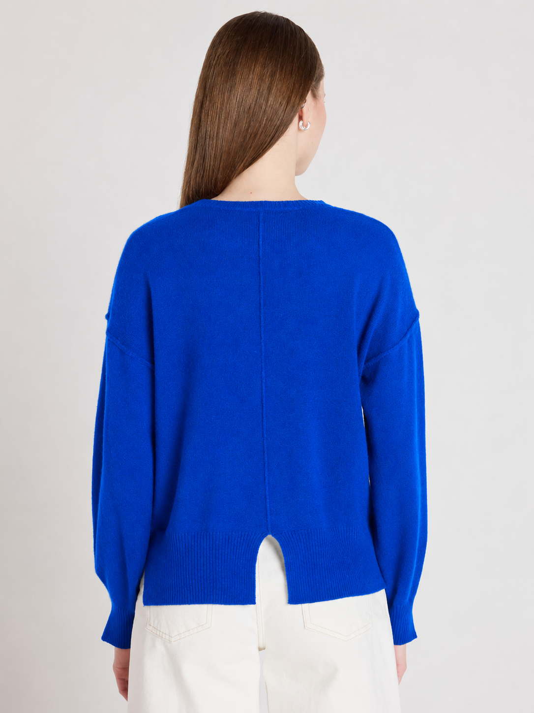 A woman viewed from the back wearing a bright royal blue knit sweater and white pants against a plain light gray studio background.