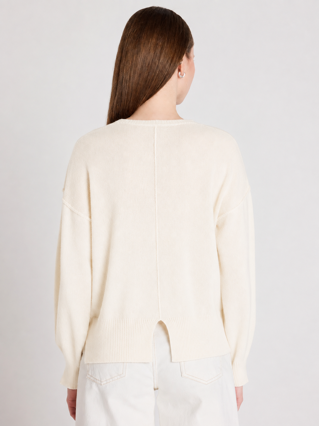 A woman viewed from the back wearing a cream-colored knit sweater and white pants against a light gray studio background.