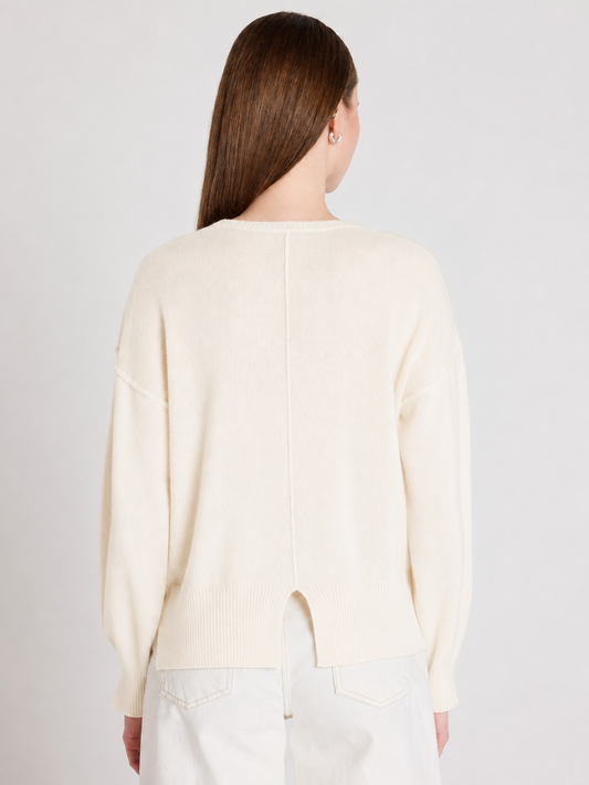 A woman viewed from the back wearing a cream-colored knit sweater and white pants against a light gray studio background.