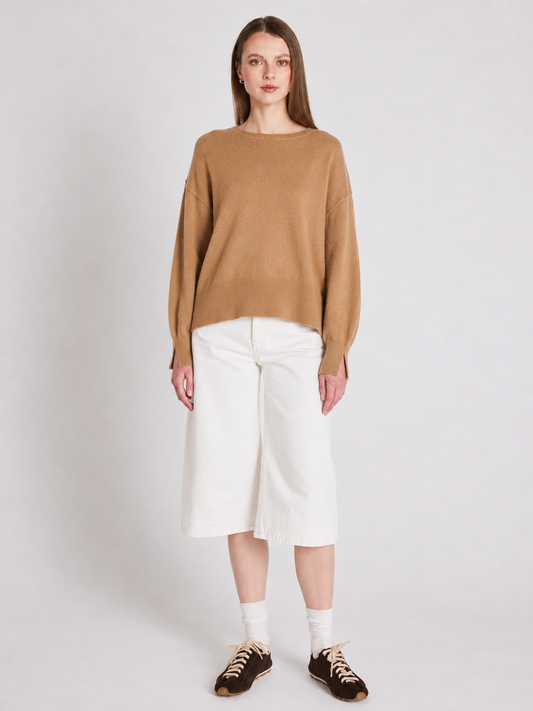 A woman stands against a light gray studio background wearing a camel-colored knit sweater, white cropped pants, white socks, and dark brown sneakers.