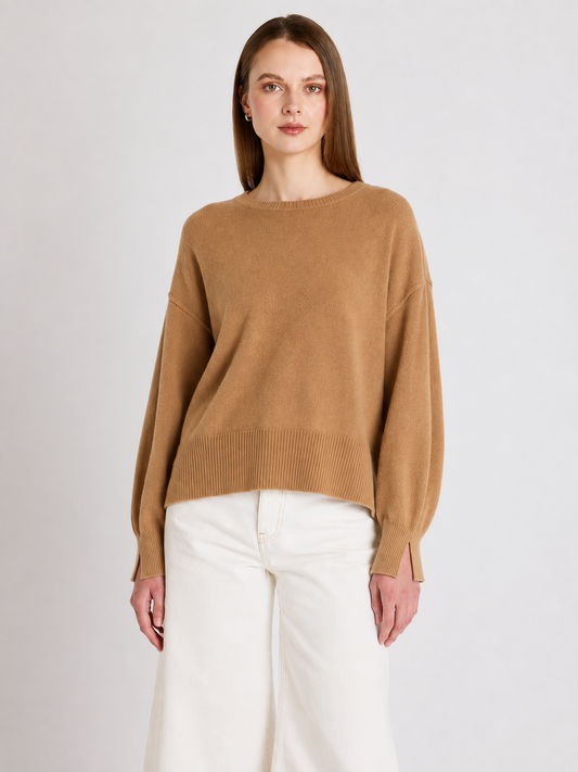 A woman modeling a camel brown knit sweater with a crew neck and dropped shoulders, paired with white wide-leg trousers, against a light gray studio background.