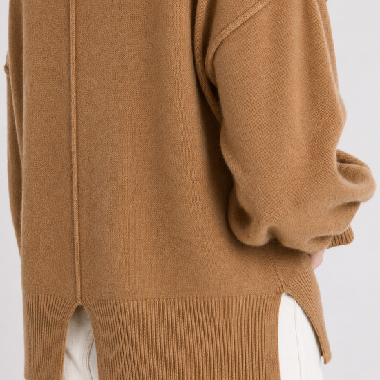 Close-up of a camel brown knit sweater worn with white pants, showing the back and side details of the garment.