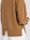 Close-up of a camel brown knit sweater worn with white pants, showing the back and side details of the garment.