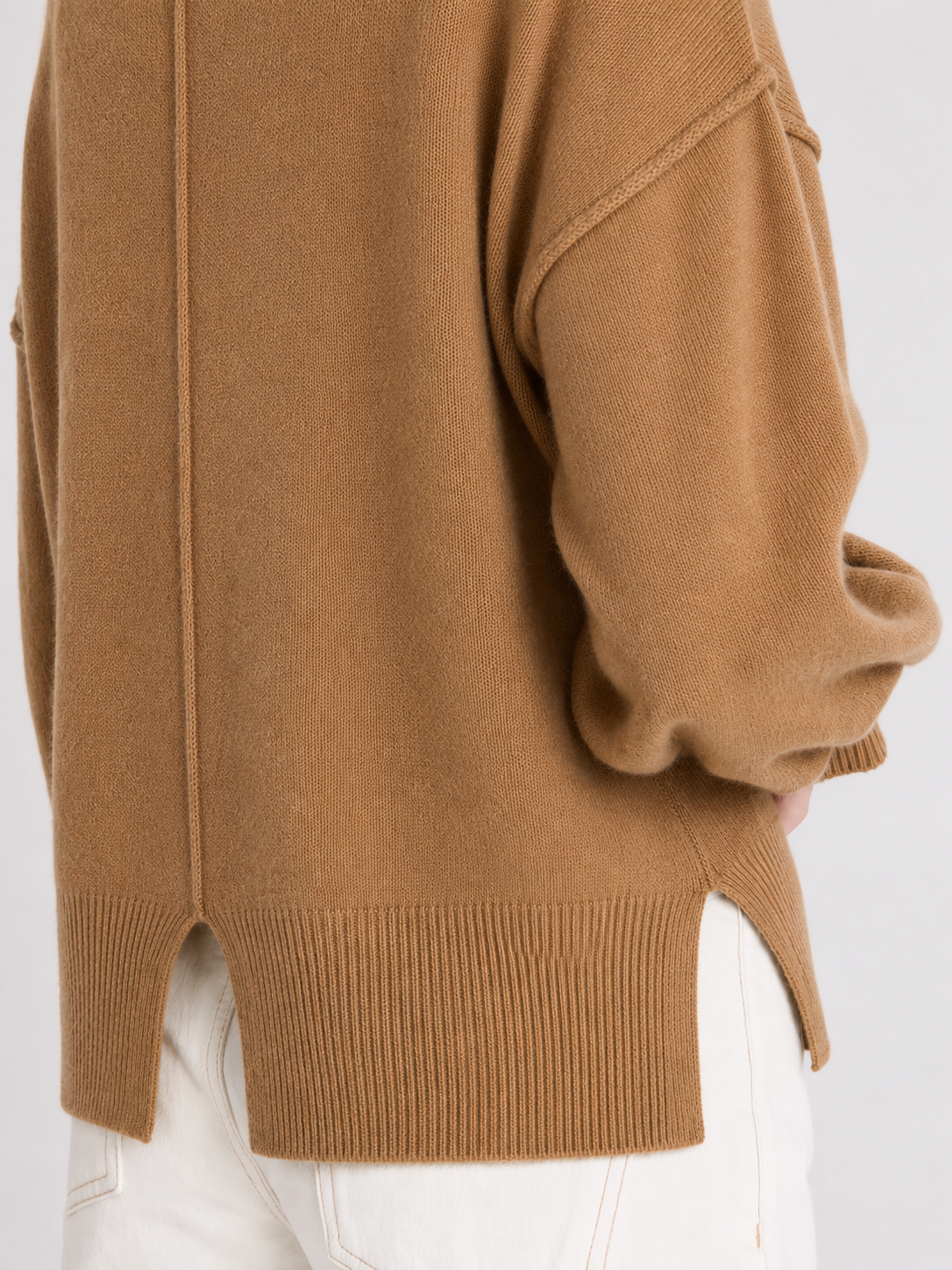 Close-up of a camel brown knit sweater worn with white pants, showing the back and side details of the garment.