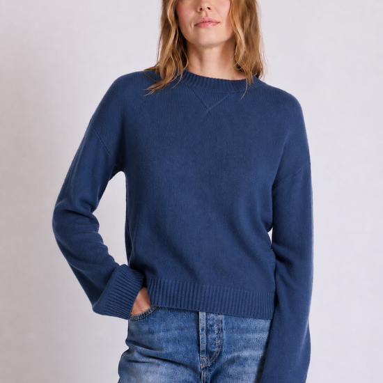 A woman wearing a navy blue crewneck knit sweater and blue jeans, standing against a light gray studio background.