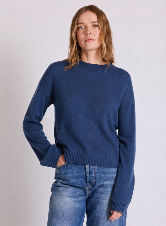 A woman wearing a navy blue crewneck knit sweater and blue jeans, standing against a light gray studio background.