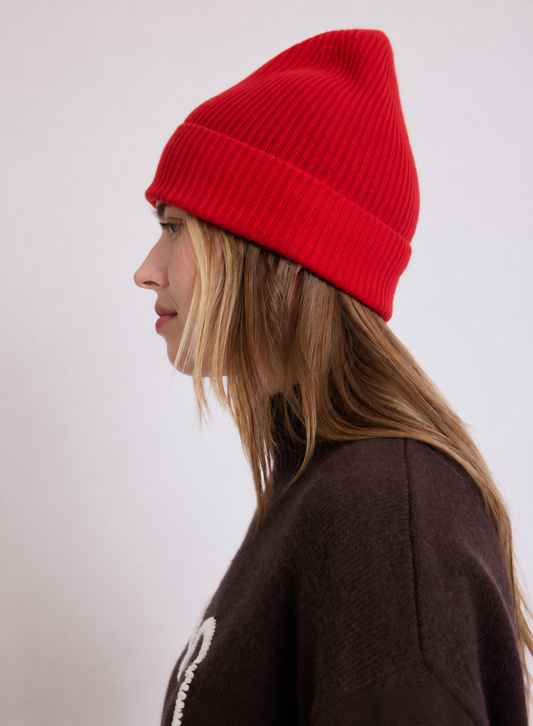 A woman in profile wearing a bright red ribbed knit beanie and a dark brown sweater, photographed against a plain light gray background.
