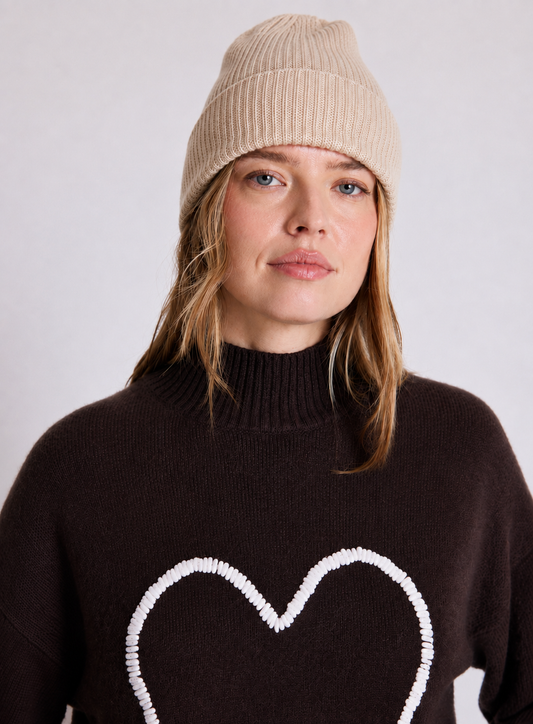 A woman wearing a beige ribbed knit beanie and a dark brown turtleneck sweater with a white beaded heart outline on the front, photographed against a light gray studio background.