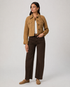 A woman modeling a cropped tan suede jacket over a white top, paired with dark brown wide-leg jeans and tan suede loafers, against a light gray studio background.