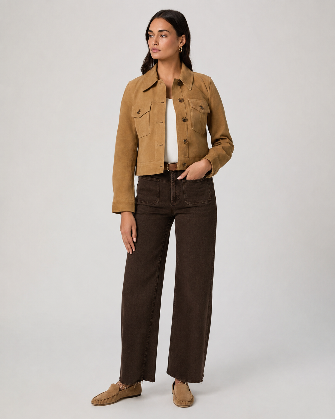 A woman modeling a cropped tan suede jacket over a white top, paired with dark brown wide-leg jeans and tan suede loafers, against a light gray studio background.