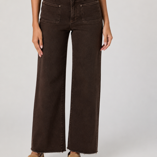 A model wearing dark brown wide-leg denim jeans with a frayed hem and tan suede slip-on shoes, shown from the waist down on a light gray studio background.