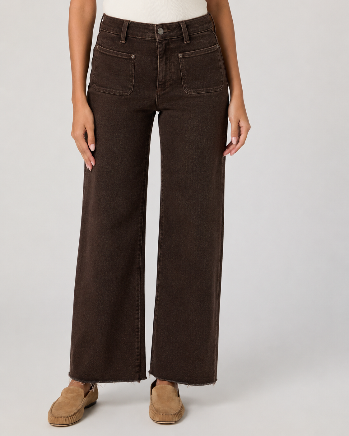 A model wearing dark brown wide-leg denim jeans with a frayed hem and tan suede slip-on shoes, shown from the waist down on a light gray studio background.