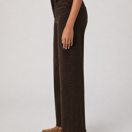 A side-profile view of a model wearing dark brown wide-leg denim jeans and tan suede moccasin loafers against a light gray studio background.