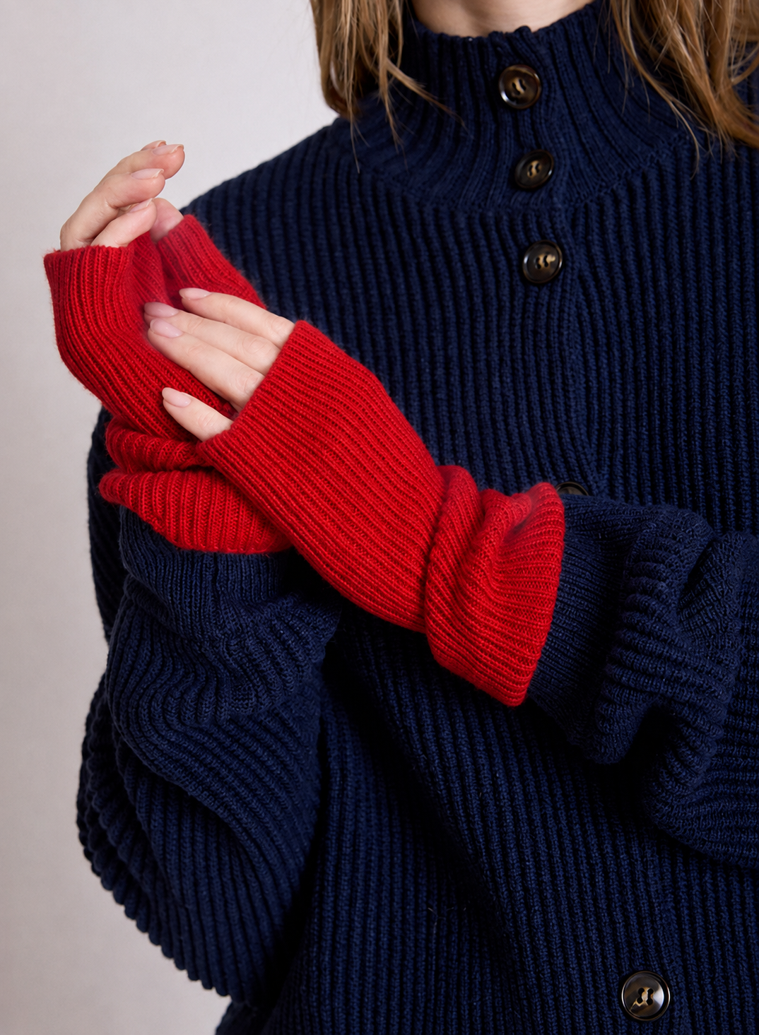 A close-up of a person wearing a navy blue ribbed knit sweater with red fingerless knit sleeves, showing the hands and upper torso against a plain light background.