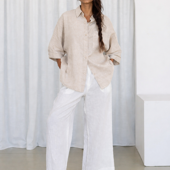 Woman wearing a beige shirt and white pants standing against a white curtain.
