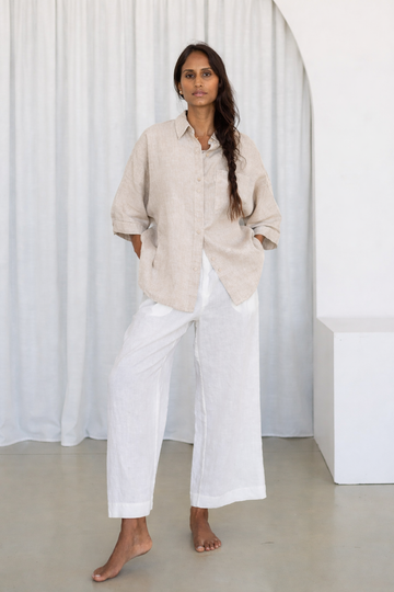 Woman wearing a beige shirt and white pants standing against a white curtain.