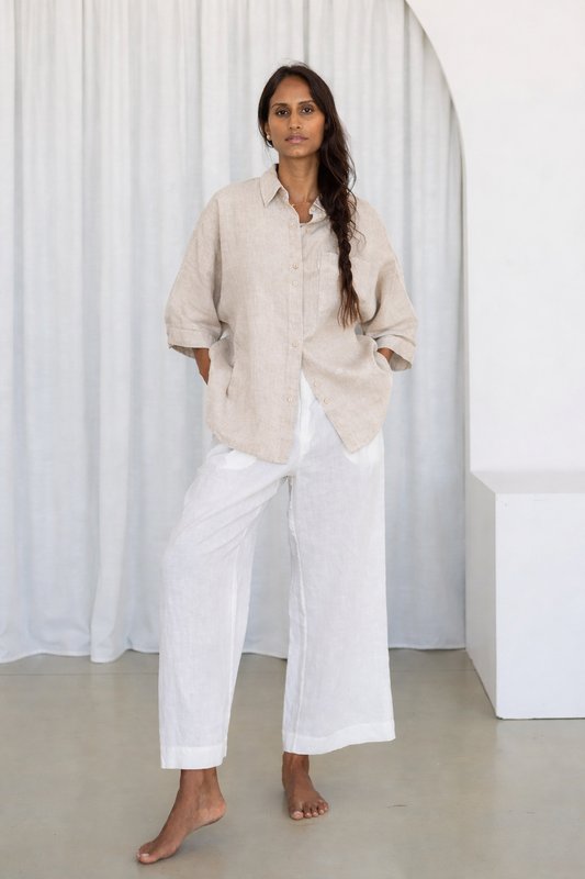 Woman wearing a beige shirt and white pants standing against a white curtain.