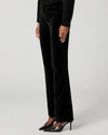 A model wearing black wide-leg velvet trousers, a black ribbed long-sleeve top, and black pointed-toe stiletto heels, shown from the waist down against a light gray studio background.
