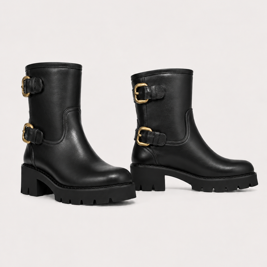 A pair of black leather ankle boots with chunky lug soles and gold-tone buckle straps on the side, displayed against a light gray studio background.