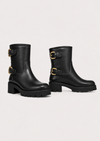 A pair of black leather ankle boots with chunky lug soles and gold-tone buckle straps on the side, displayed against a light gray studio background.