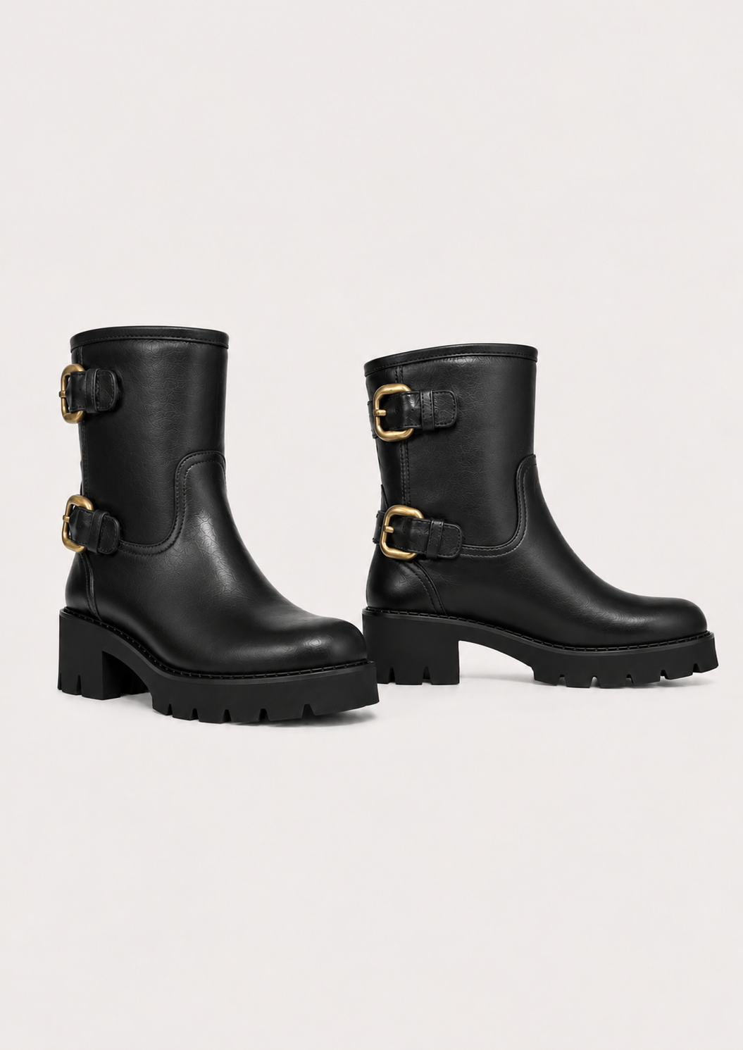 A pair of black leather ankle boots with chunky lug soles and gold-tone buckle straps on the side, displayed against a light gray studio background.